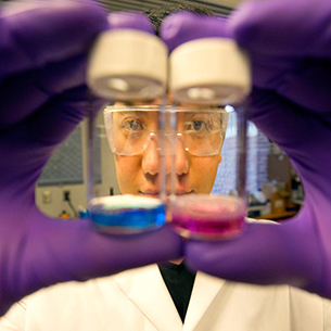 Student face behind two purple gloved hands holding a blue liquid and a pink liquid